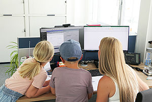 Zwei Kinder und ein Erwachsener betrachten gemeinsam zwei Computerbildschirme in einem Büro.