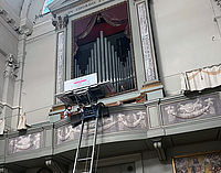 Ein Aufzug transportiert eine Orgelpfeife in einer Kirche mit hohen Wänden und großen Bögen.