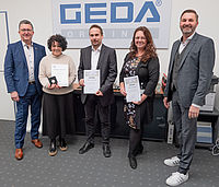 Fünf Personen stehen nebeneinander, drei halten Auszeichnungen vor einem GEDA Schild.