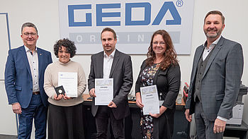 Fünf Personen stehen nebeneinander, drei halten Auszeichnungen vor einem GEDA Schild.