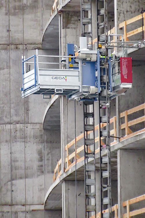 Ein Bauaufzug bewegt sich an einer Betonstruktur mit abgerundeten Balkonen aufwärts.
