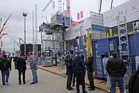 Menschen stehen und unterhalten sich vor großen Baumaschinen und Ausstellungskonstruktionen auf einer Messe im Freien.