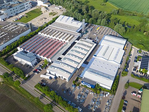 Industriekomplex mit langen Hallen steht inmitten grüner Landschaft, umgeben von Straßen und Bäumen.