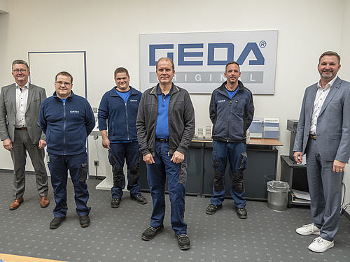 Sechs Männer stehen lächelnd vor einem GEDA-Original-Logo in einem Büro.