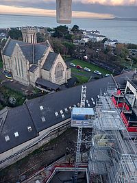 Ein Bauaufzug bewegt sich vor einem hohen Gebäude mit Gerüst, während im Hintergrund eine Kirche und einige Autos in einer Wohngegend nahe dem Meer sichtbar sind.