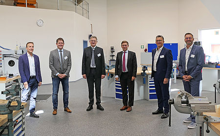 Sechs Männer stehen lächelnd zusammen in einer Werkstatt mit Werkbänken und Maschinen im Hintergrund.