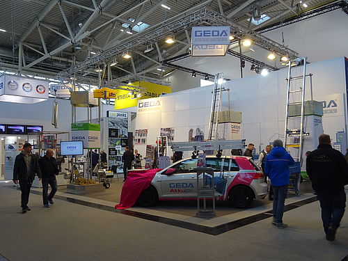 Ein Messestand zeigt Hebe- und Fördergeräte von GEDA, umgeben von Besuchern in einer Ausstellungshalle.