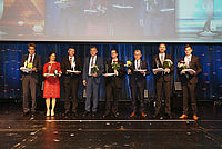 Sieben Personen in formeller Kleidung halten Blumensträuße und Dokumentenmappen.