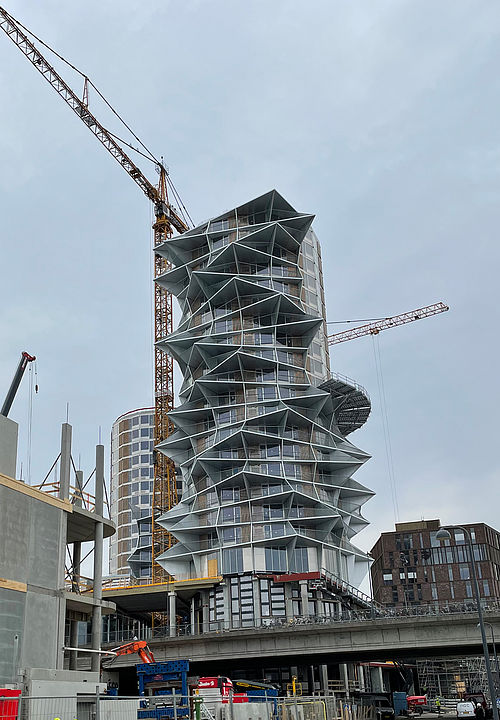 Ein modernes, mehrstöckiges Gebäude mit markanter, geometrischer Fassade wird von zwei Baukränen umgeben.