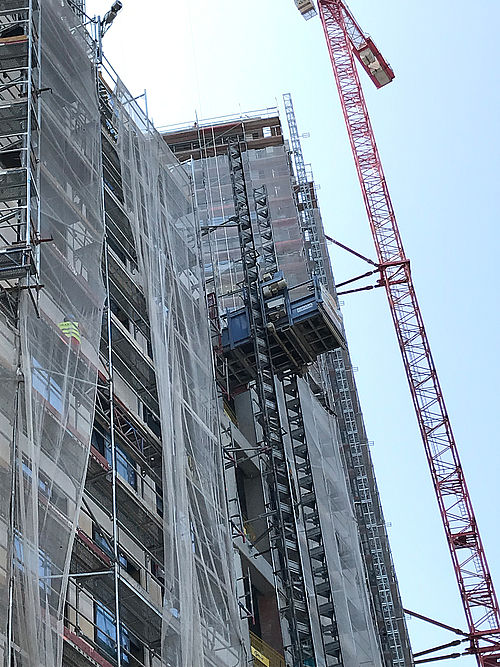 Gerüstaufzug transportiert Materialien; neben einem Hochhaus mit Schutzplanen und einem Kran.