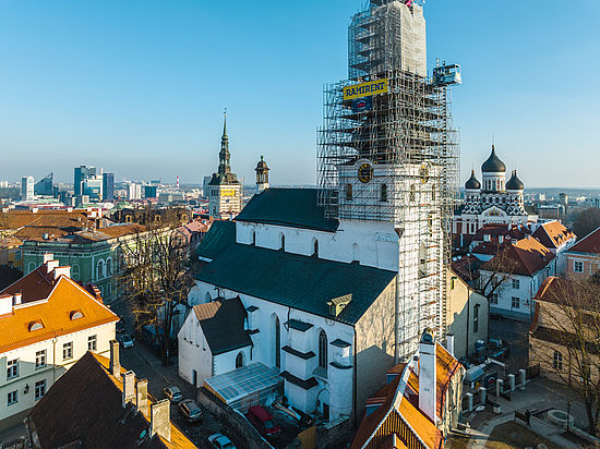Ein Kirchturm ist vollständig in ein Baugerüst eingehüllt, im Hintergrund die Skyline einer Stadt.