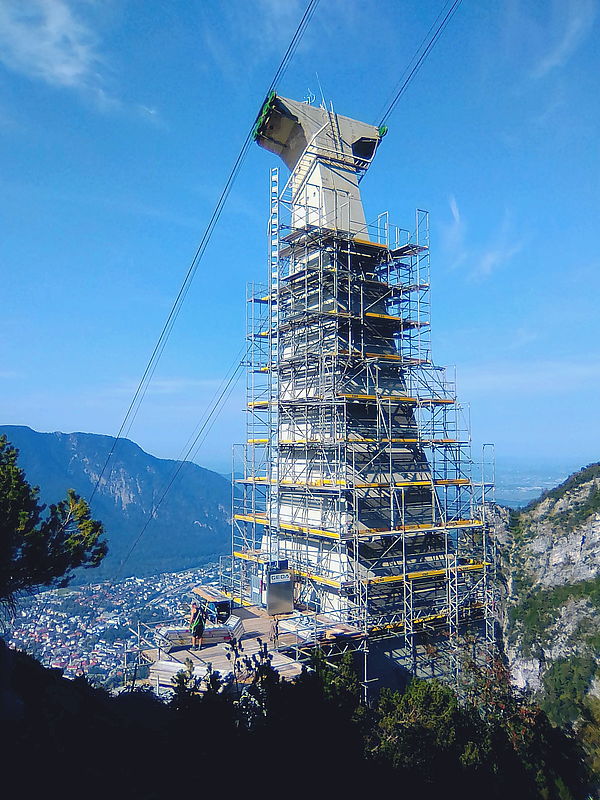 Ein mit Gerüst umgebenes Seilbahnstütze wird gewartet, im Hintergrund erstreckt sich eine Berglandschaft und eine Stadt im Tal.
