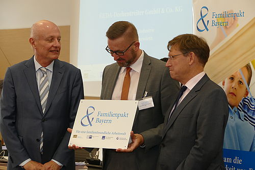 Drei Männer stehen eng zusammen; der mittlere hält ein Schild mit der Aufschrift „Familienpakt Bayern - Für eine familienfreundliche Arbeitswelt“.