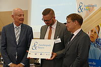 Drei Männer stehen eng zusammen; der mittlere hält ein Schild mit der Aufschrift „Familienpakt Bayern - Für eine familienfreundliche Arbeitswelt“.