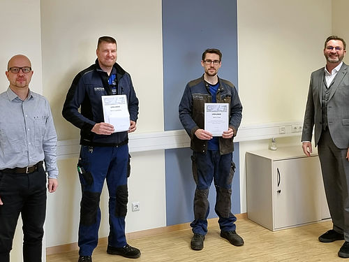Vier Männer stehen in einem hellen Raum, zwei halten Zertifikate mit der Aufschrift „Fachkraft für festgelegte Tätigkeiten“.