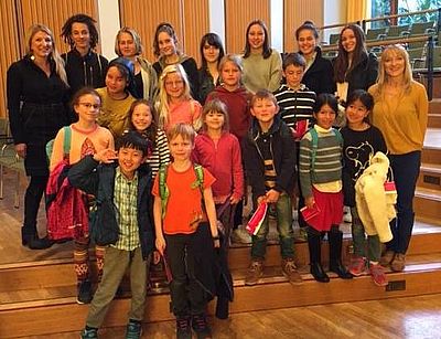 Eine Gruppe von Kindern und Erwachsenen steht auf gestuften Holztribünen in einem hellen Raum mit Vorhängen und Fenstern.