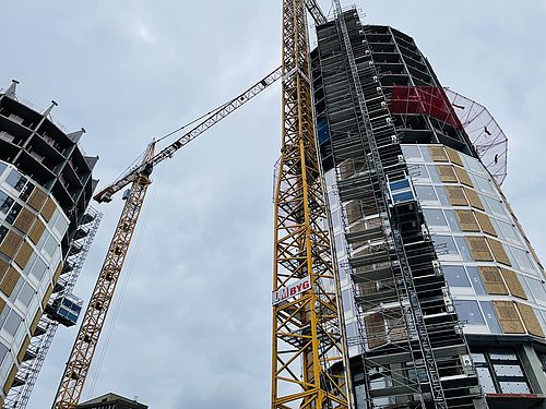 Zwei gelbe Kräne heben Baumaterialien neben zwei im Bau befindlichen gläsernen Hochhaustürmen.