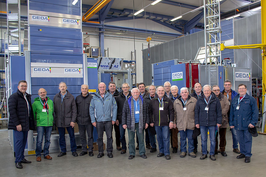 Eine Gruppe von Männern steht in einer Fabrikhalle vor Maschinen mit der Aufschrift "GEDA".