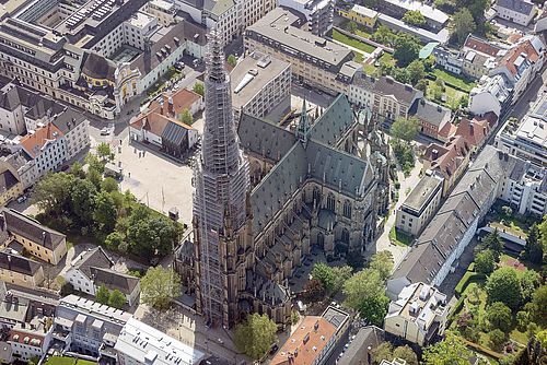 Eine große gotische eingerüstete Kathedrale mit einem hohen Turm steht im Zentrum einer Stadt, umgeben von Gebäuden und grünen Bäumen.