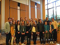 Eine Gruppe von Jugendlichen und zwei Erwachsenen stehen zusammen, lächelnd, in einem Raum mit hölzernen Wänden und großen Fenstern im Hintergrund.