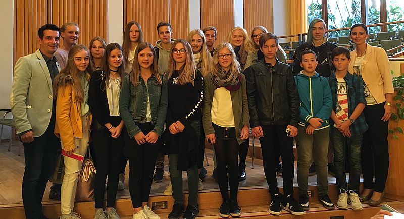 Eine Gruppe von Jugendlichen und zwei Erwachsenen stehen zusammen, lächelnd, in einem Raum mit hölzernen Wänden und großen Fenstern im Hintergrund.