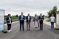 Sechs Personen halten ein blaues Band bei einer Einweihungszeremonie auf einer asphaltierten Straße unter bewölktem Himmel.