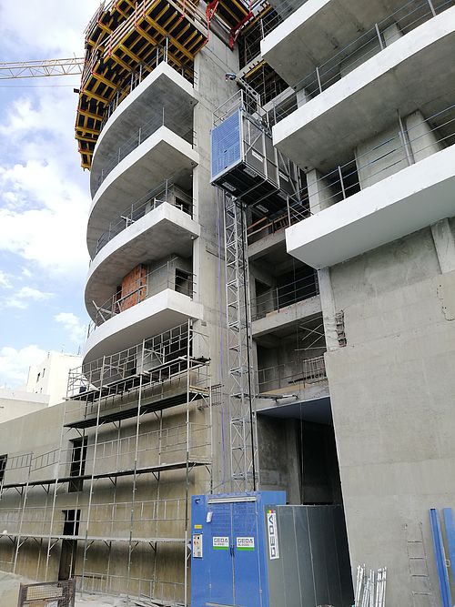 Ein Rohbau eines mehrstöckigen Gebäudes steht mit sichtbaren Betondecken, Gerüsten und einem Bauaufzug.
