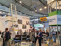 Messebesucher stehen und unterhalten sich in einem Ausstellungsbereich mit verschiedenen Hebegeräten und Infotafeln der Marke GEDA.
