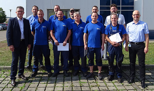 Eine Gruppe von elf Männern posiert lächelnd für ein Foto auf einem gepflasterten Platz vor einem großen Industriegebäude.