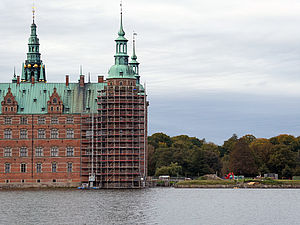 Ein Schloss mit grünen Dächern, das einen Baugerüst an einer Turmseite trägt, steht am Wasser, umgeben von Bäumen und bedecktem Himmel.