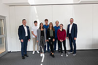 Eine Gruppe von acht Personen steht lächelnd auf einem grauen Teppichboden in einem Raum mit weißen Wänden.