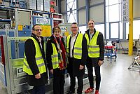 Vier Personen tragen gelbe Sicherheitswesten und stehen lächelnd vor einer GEDA 1200 Z/ZP Hebeanlage in einer Fabrikhalle mit großen Fenstern und industrieller Ausstattung.