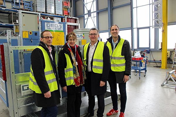 Vier Personen tragen gelbe Sicherheitswesten und stehen lächelnd vor einer GEDA 1200 Z/ZP Hebeanlage in einer Fabrikhalle mit großen Fenstern und industrieller Ausstattung.