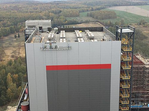 Ein großes Gebäude mit grauer und roter Fassade steht in einer bewaldeten Landschaft.