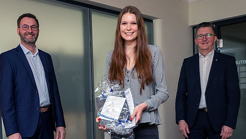 Eine Frau hält ein Geschenkpaket, während zwei Männer lächelnd danebenstehen.