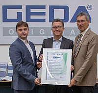 Drei Männer halten ein Zertifikat mit der Aufschrift „ZERTIFIKAT GEDA ISO 45001:2018“ in einem Büro, vor einem großen „GEDA“ Logo an der Wand.