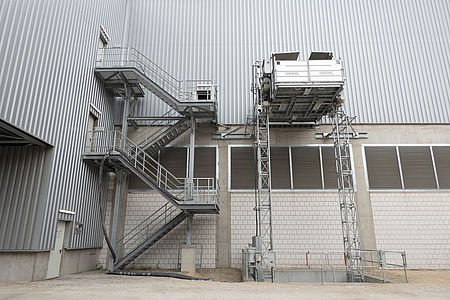 Eine metallene Außentreppe führt mehrstöckig entlang einer grauen Industriehallenwand, daneben eine Hebebühne.