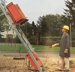Eine rote Hebebühne transportiert Baumaterial schräg nach oben, daneben ein Mann mit gelbem Sicherheitshelm.