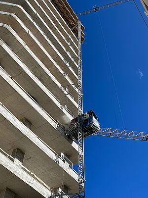 Ein Baukran hebt Baumaterialien an einem mehrstöckigen Rohbau empor.