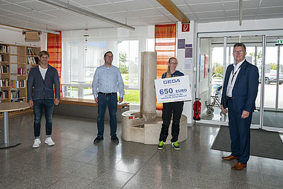 Vier Personen stehen in einem Foyer, einer hält ein großes Schild mit der Aufschrift "GEDA 650 EURO Sportverein".