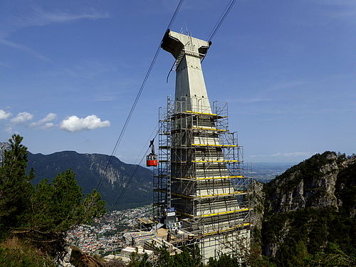 Ein hoher Turm wird genoviert, während eine rote Seilbahnkabine vorbeifährt.