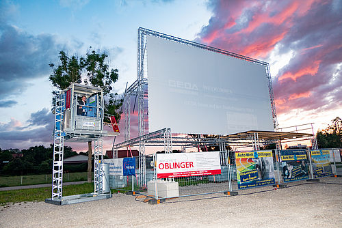 Große Leinwand, umgeben von einem Bauzaun mit Werbebannern und einer Personenhebebühne daneben.