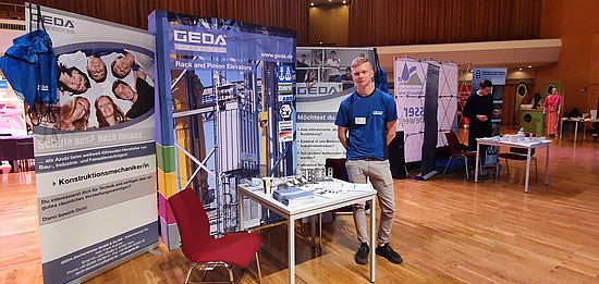 Ein Mann steht lächelnd an einem Messestand mit Broschüren und einem Roll-up von GEDA, das Aufzüge illustriert.