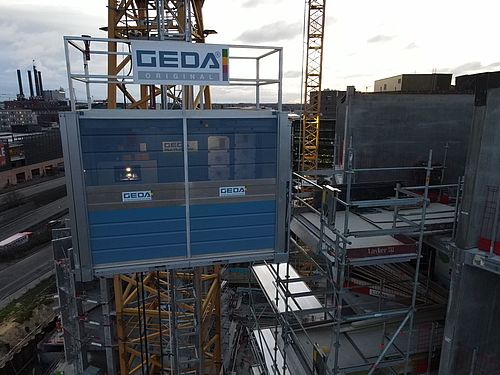 Ein Bauaufzug von GEDA bewegt sich entlang eines Gerüsts vor einem Kran.