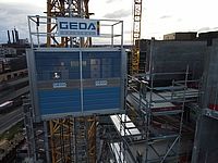 Ein Bauaufzug von GEDA bewegt sich entlang eines Gerüsts vor einem Kran.