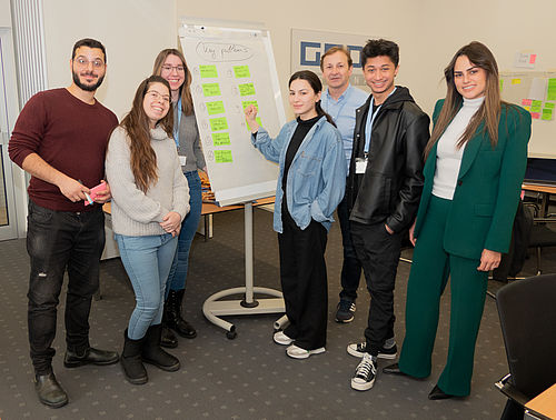 Eine Gruppe von sechs jungen Erwachsenen steht lächelnd um ein Flipchart mit darauf geklebten grünen Notizzetteln in einem Konferenzraum.