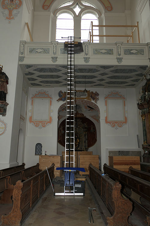 Eine lange, aufrechte Leiter führt zu einer erhöhten Galerie in einer Kirche, umgeben von verzierten Wänden und Bänken.