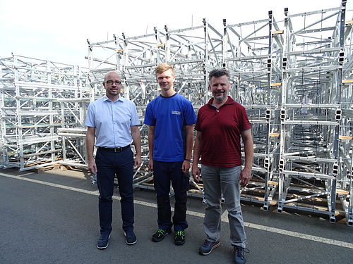 Drei Männer stehen auf einer asphaltierten Fläche, umgeben von großen Stapeln metallischer Gerüste.