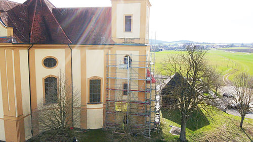Gerüst steht an der Wand einer gelben Kirche, während Bäume und eine weite, grüne Landschaft im Hintergrund sichtbar sind.