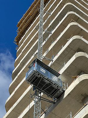 Ein Bauaufzug transportiert Baumaterialien entlang eines halb fertigen Hochhauskomplexes in die Höhe; blauer Himmel und teilweise Wolken bilden den Hintergrund.